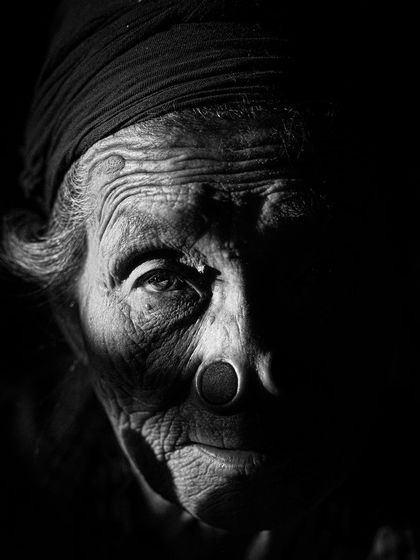 A low-key, black and white portrait of an Apatani woman. The dramatic lighting focuses on one side of her face, emphasizing her traditional nose plug and the wisdom in her eyes.