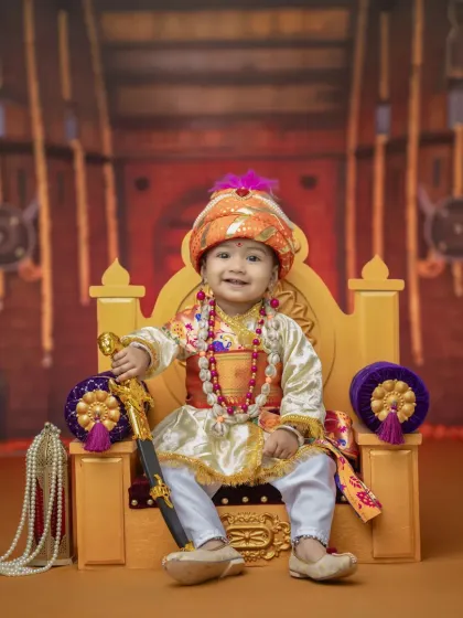 A regal portrait of a baby boy in a Bal Shivba costume, holding a toy sword. The rich fabrics of the costume and the ornate throne create a picture of miniature royalty.