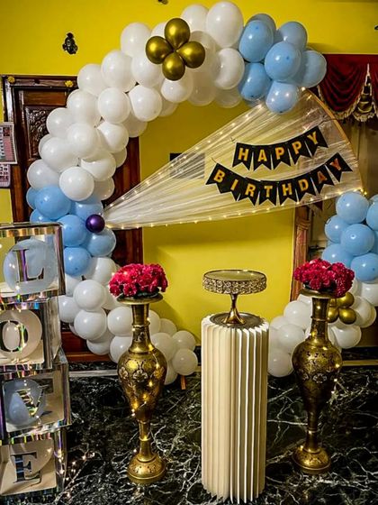 An elegant home setup featuring a white and light blue balloon arch. The decor includes tall brass vases with red roses, a central pedestal for the cake, and iridescent 'LOVE' blocks, creating a sophisticated party atmosphere.