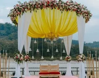 This mandap features a canopy of bright yellow fabric with a floral border. Hanging bells and a central chandelier add a traditional and elegant touch to the riverside ceremony setup.