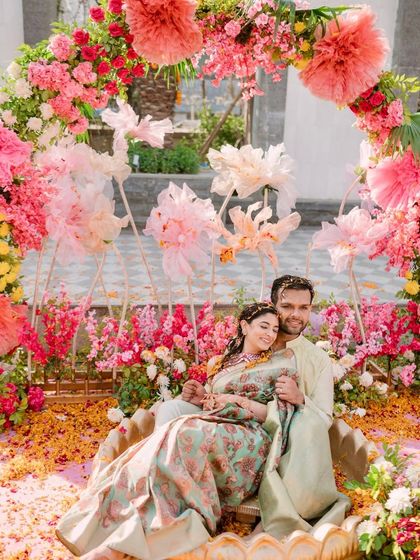 A dreamy Haldi seating arrangement where the couple is nestled in a giant brass urli, surrounded by a floral arch of pink and yellow blooms and whimsical paper flowers.
