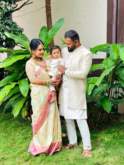 The family of three posing together. The father holds the baby, showing off their perfectly coordinated cream-colored traditional wear.