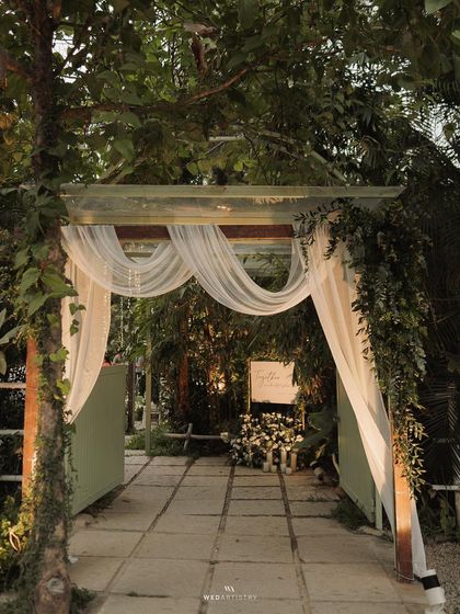 The entrance to an evening wedding reception, elegantly draped in white fabric and greenery. The welcome sign invites guests into a magical, warmly lit space.