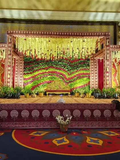 A beautiful example of a traditional stage for a wedding ceremony. The design features a rich floral backdrop using red and green flowers, framed by structures draped in classic wedding colors and patterns.