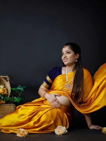 A beautiful studio portrait capturing the elegance of a mother-to-be in a flowing yellow saree. The dark background and simple props create a timeless and artistic feel.