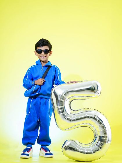 A full-length shot of a boy celebrating his fifth birthday, holding a giant silver balloon against a bright yellow background.