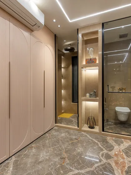 This spacious bathroom combines a walk-in closet with the main area. The design features soft pink, fluted wardrobe doors, marble flooring, and a glass-enclosed shower.