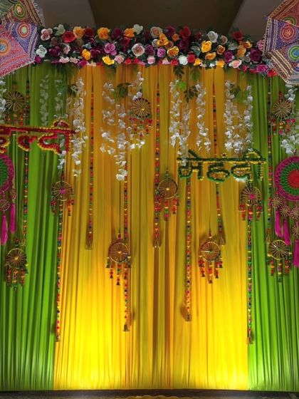 A beautiful Haldi and Mehandi backdrop with lime green and yellow drapes. The decor includes hanging dreamcatchers, tassels, and Rajasthani umbrellas for a bohemian and festive look.