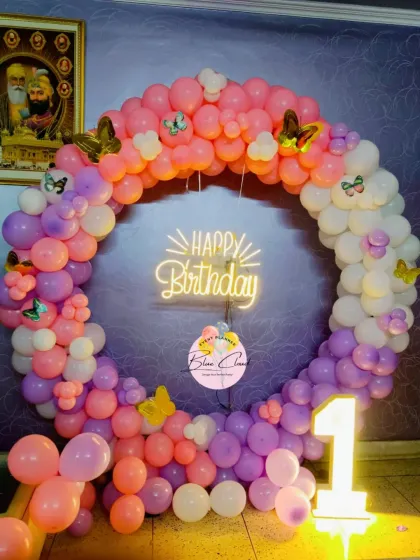 A beautiful butterfly-themed first birthday with a full ring of pastel pink, purple, and white balloons. A central 'Happy Birthday' neon sign and a lit-up number '1' make it picture-perfect.