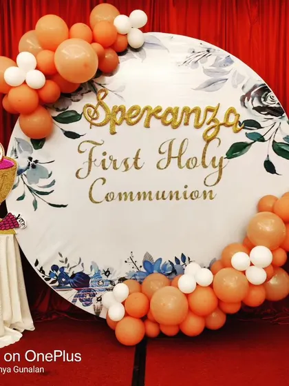 A close-up of the custom circular backdrop for a First Holy Communion. The design features delicate floral graphics, elegant gold lettering, and a tasteful balloon garland in peach and white tones.