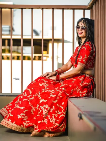 A modern bride takes a moment. Dressed in a stunning red lehenga and sunglasses, she relaxes against a modern slatted wall, bathed in golden hour light.