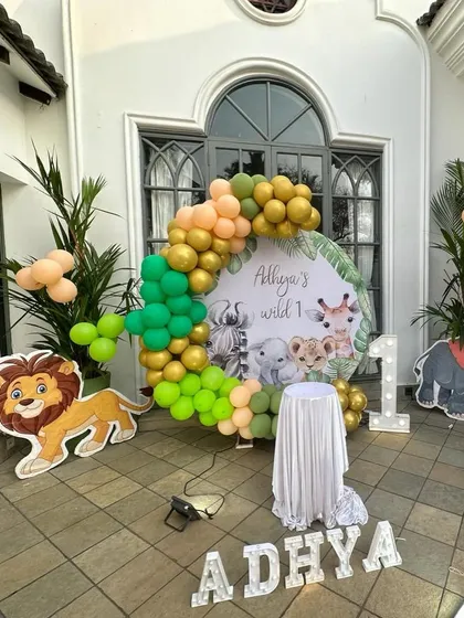 A 'Wild One' first birthday party with a vibrant jungle safari theme. This outdoor setup includes a custom backdrop with cute animal illustrations, a balloon arch in green, gold, and peach, and character cutouts like a lion and elephant.
