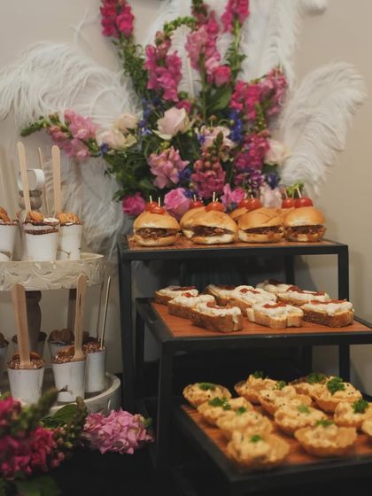 A beautiful display of appetizers for a luxury event. The spread includes mini sliders, bruschetta, and other savory tarts, all complemented by a stunning floral arrangement.