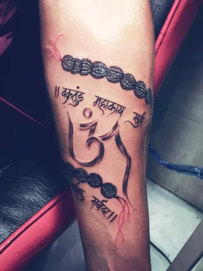 An Om symbol tattoo on the forearm, designed with a Rudraksha mala. The piece is wrapped around the arm, incorporating a Ganesha mantra in Sanskrit script.