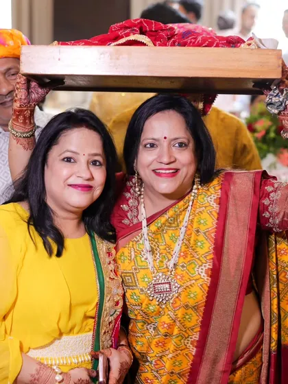 Two smiling women carrying a decorative tray on their heads as part of a traditional wedding ritual.