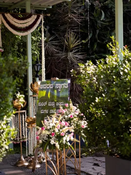We create welcoming entrances that set the perfect tone for your celebration. This setup for a home function features a custom welcome sign in Kannada, traditional brass lamps, and lush floral arrangements.