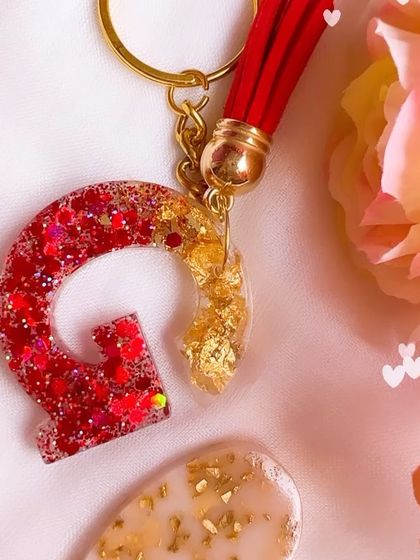 A detailed shot of the 'G' initial keychain, showing the blend of red glitter and gold leaf, attached to a keychain ring with a matching tassel.