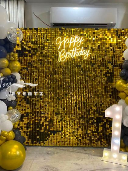 I designed this glamorous and modern birthday backdrop using a gold shimmer wall. The setup is framed by an organic balloon garland in black, white, and gold, and features a 'Happy Birthday' neon sign and a large marquee number '1' to mark the special occasion.