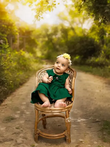 A beautiful outdoor portrait of a baby girl in a green dress, seated on a small wicker chair on a country lane. The soft, natural light is perfect for baby photography.
