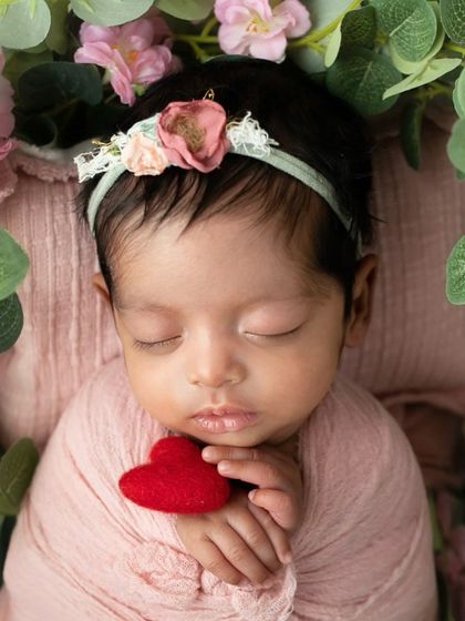 A newborn girl holds a tiny red heart, surrounded by soft pink flowers. A sweet and simple portrait that is full of love.
