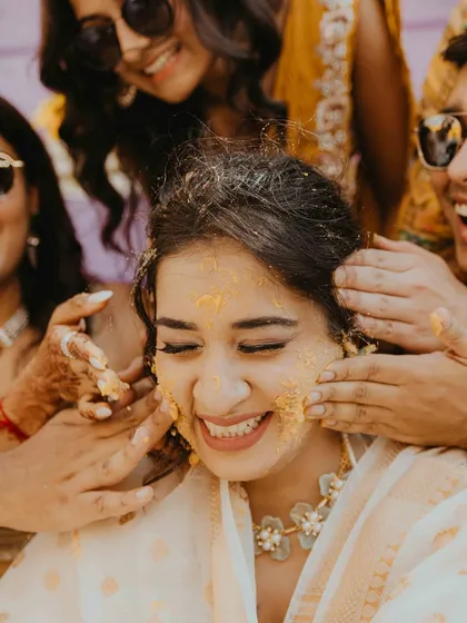 The bride closes her eyes and smiles as her friends and family lovingly apply Haldi paste. A beautiful, candid moment of tradition and affection.