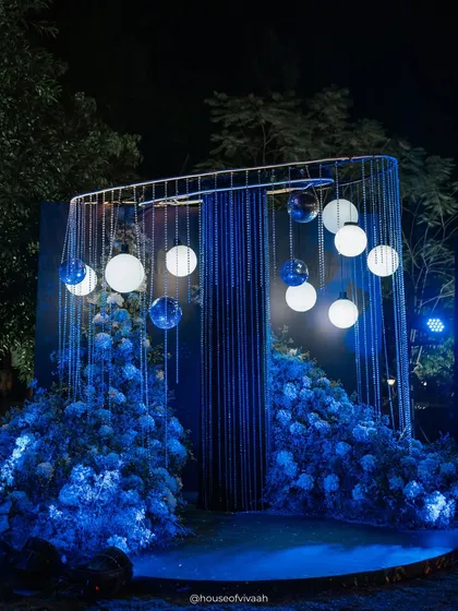 A stunning photo installation for a "sky full of stars" Sangeet, featuring suspended glowing orbs within a cage-like structure, surrounded by clouds of blue hydrangeas.