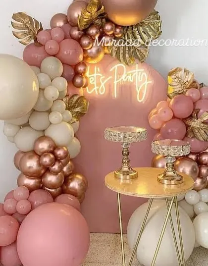 A chic arrangement featuring a pink backdrop with a 'Let's Party' neon sign. The organic balloon garland in shades of pink, cream, and rose gold is accented with gold palm leaves for a tropical glam look.