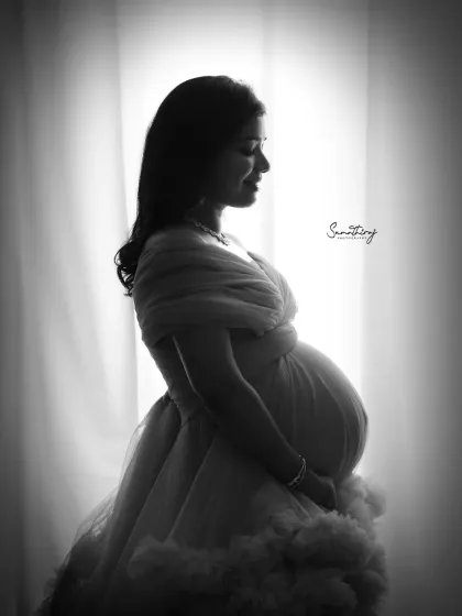 A beautiful black and white silhouette of a mother-to-be in a flowing gown. The soft light from the window creates a dreamy and ethereal effect, highlighting her graceful form.