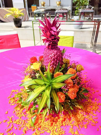 A creative and colorful table centerpiece for a tropical-themed Haldi, featuring a painted pink pineapple and bright marigolds.