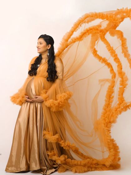 A graceful studio portrait of a mother-to-be in a golden gown with flowing orange wings. This creative setup gives the image an ethereal, angelic quality.