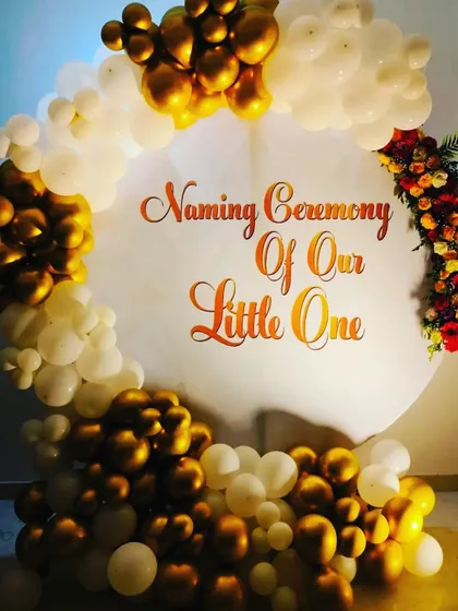 An elegant naming ceremony backdrop with a simple and clean design. The focus is on the "Naming Ceremony Of Our Little One" text, framed by a beautiful arch of gold and white balloons with a touch of floral arrangement.
