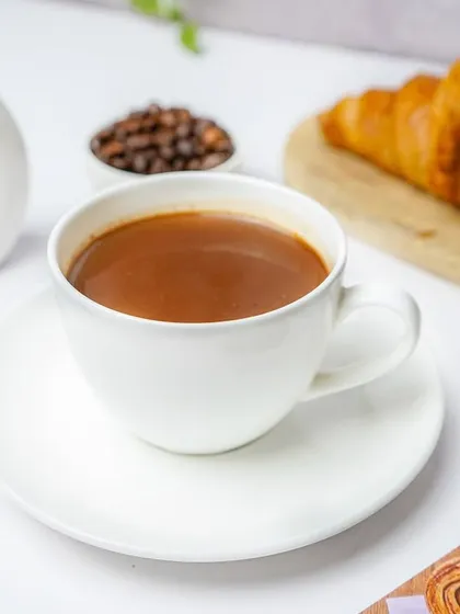 A cup of coffee in a bright, clean setting with a white vase and a croissant. This lifestyle shot is perfect for a cafe's website or Instagram page, selling an experience.