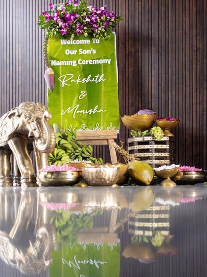 A personalized welcome sign for the naming ceremony, placed on a banana leaf and easel. This is complemented by a brass elephant statue and bowls of fresh flower petals for guests.
