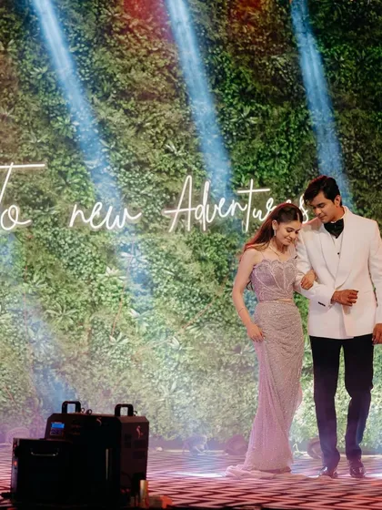 "To new adventures." The couple walks into their celebration, framed by a neon sign and a lush green wall. This image blends modern decor with the classic elegance of their attire.