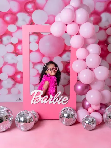 A vibrant 'Barbie' themed photoshoot for a stylish toddler. She poses confidently in a pink frame against a wall of pink balloons and disco balls, complete with a feather boa and sunglasses.