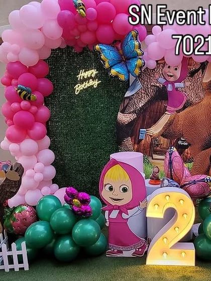 A charming Masha and the Bear themed outdoor party. This setup includes a green grass backdrop, character cutouts, and a pink balloon arch, perfect for a garden celebration.