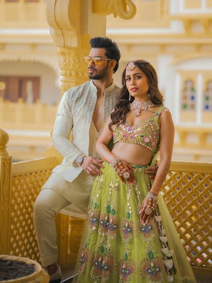 A fashionable couple portrait from a Mehendi or Haldi event, set against ornate palace architecture.