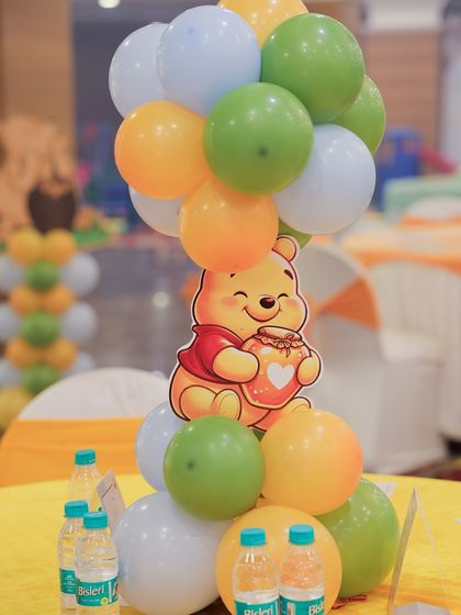 A cute Winnie the Pooh centerpiece for guest tables. Pooh Bear holds a honey pot while sitting on a cloud of orange, green, and white balloons.