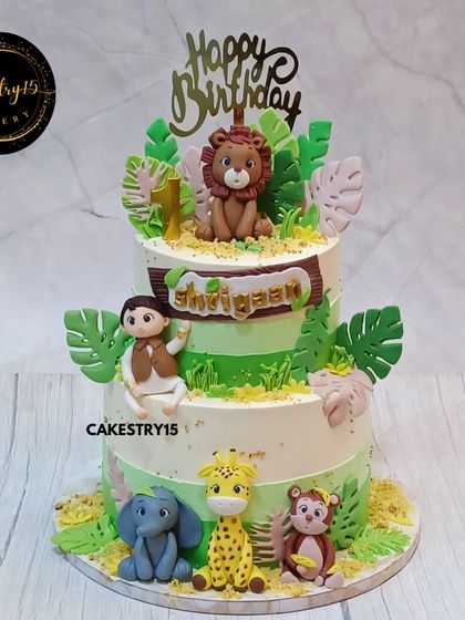 A vibrant two-tier jungle-themed cake for a first birthday. This 3kg mixed fruit cake features a boy figurine and various fondant animals, including a lion, giraffe, and elephant.