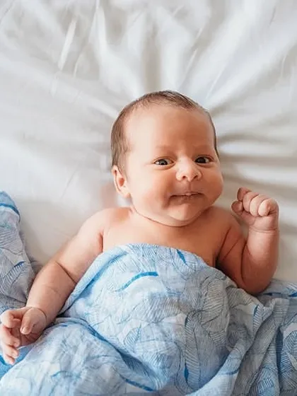 A happy and awake newborn peeking out from under a blue blanket. Capturing eye contact and early smiles is a special part of our baby photoshoots.