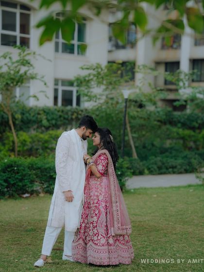 A romantic outdoor portrait of the couple. The bride's pink lehenga and the groom's white sherwani look beautiful against the lush green background.