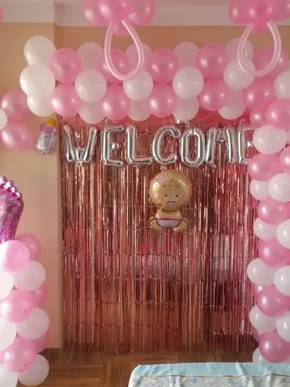 A 'Welcome Baby' girl decoration in pink and white, with a balloon arch over a shimmer curtain.
