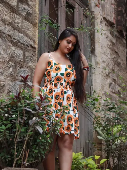 A candid-style shot of the model in the sunflower dress, looking down, set against a stone building.