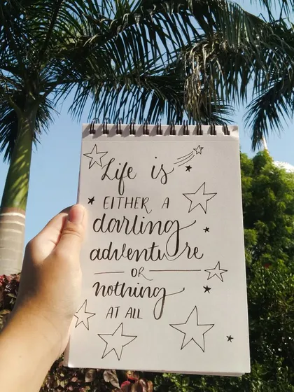 A simple calligraphy piece held up against the sky: "Life is either a daring adventure or nothing at all."