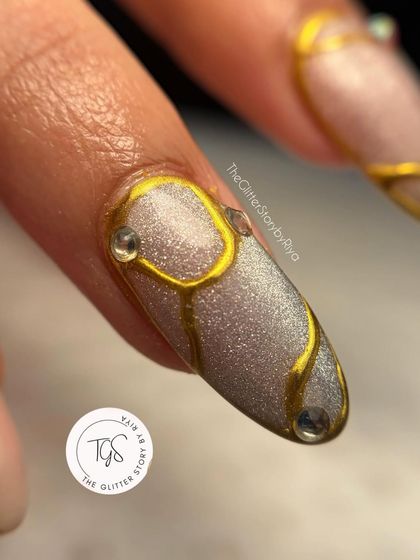 An extreme close-up of a nail with flowing, sculpted gold lines over a glittery base. Tiny clear crystals add a subtle touch of sparkle to the design.
