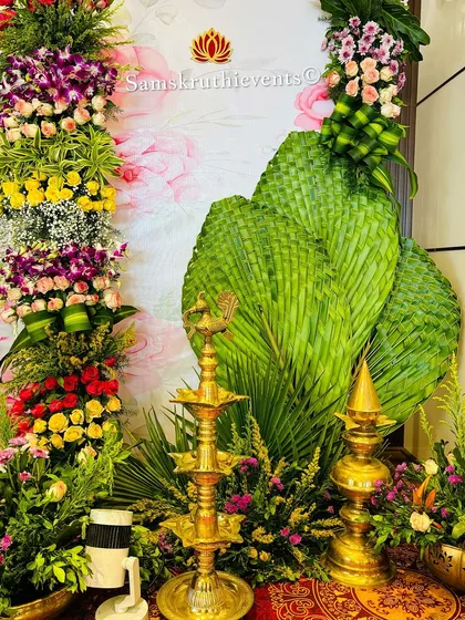 A close-up of a corner decoration, showing a traditional brass lamp placed amidst fresh flowers and woven coconut leaves. These elements are key to creating an authentic South Indian vibe.