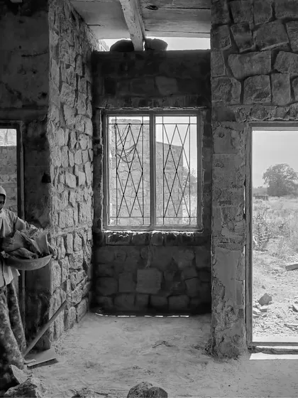 An interior view during construction. The rugged stone walls are in place, and a worker clears debris. The openings for doors and windows already frame the landscape outside.