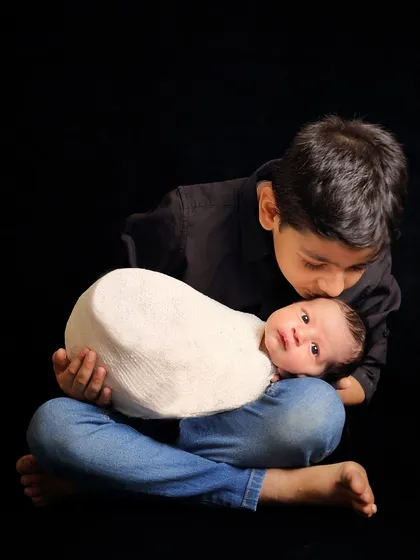 A simple yet powerful moment of a big brother giving his newborn sibling a gentle kiss on the forehead. The dark background makes this tender interaction the sole focus.