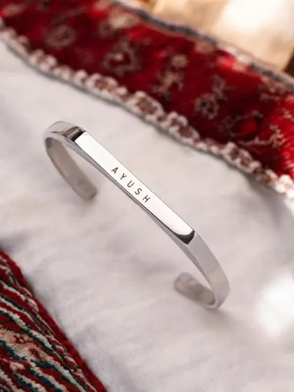 A silver cuff engraved with 'AYUSH', displayed on a white cloth with a traditional red patterned border, showing its versatility across different styles.
