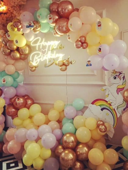 This unicorn theme photo booth uses a pastel balloon arch and a 'Happy Birthday' neon sign. A unicorn cutout on the side adds to the magical feel of the setup.